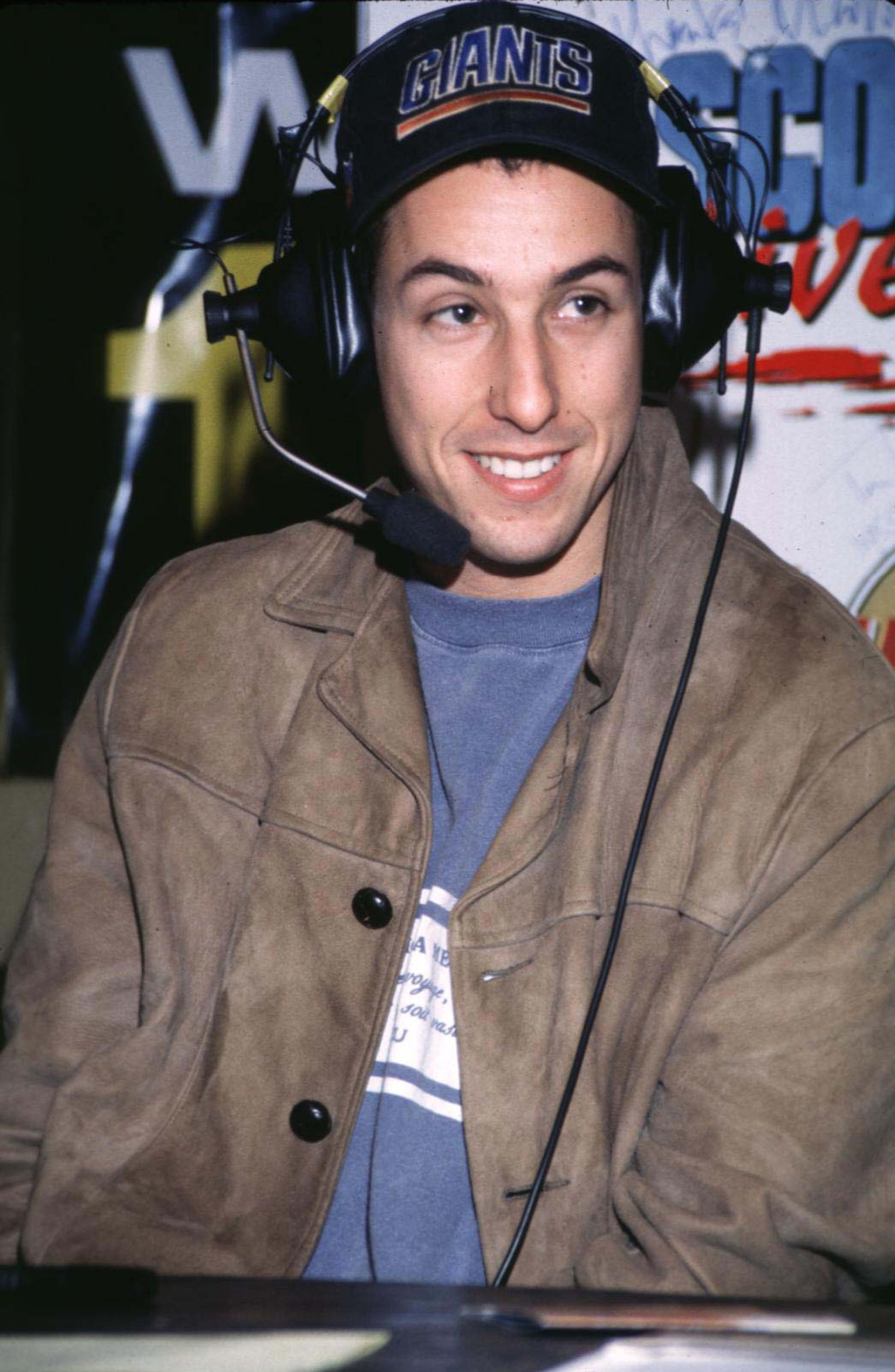 Adam Sandler wearing a Giants cap and headset, smiling while seated at a table. He is dressed in a casual jacket and a T-shirt