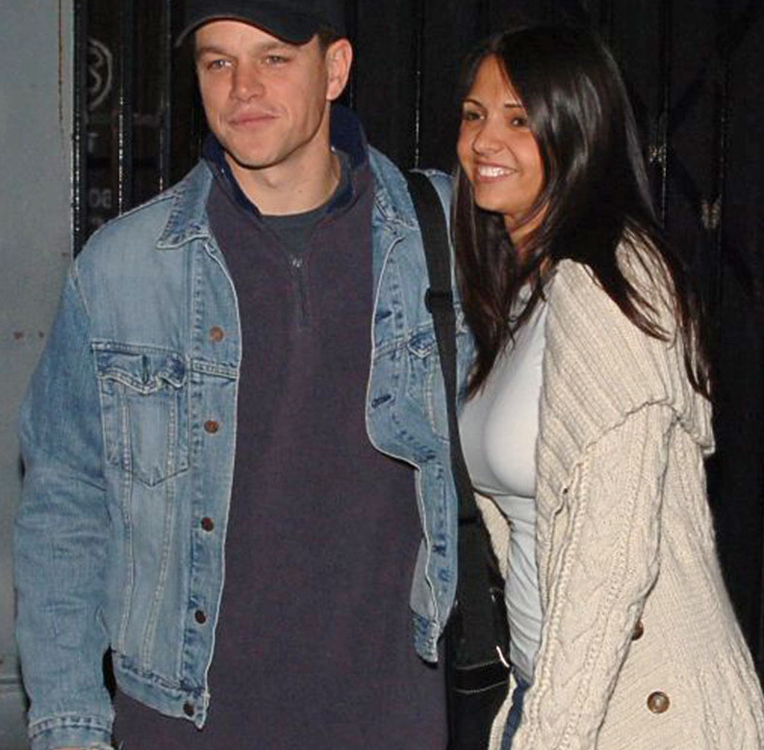 Matt Damon wearing a denim jacket and a Red Sox cap, standing next to Luciana Barroso in a knit cardigan. They are posing and smiling outdoors