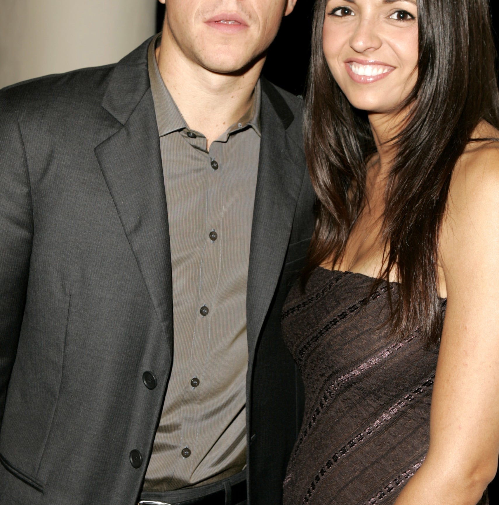 Matt Damon, in a gray suit and brown shirt, stands next to Luciana Barroso, wearing a strapless, dark-patterned dress, smiling at an event