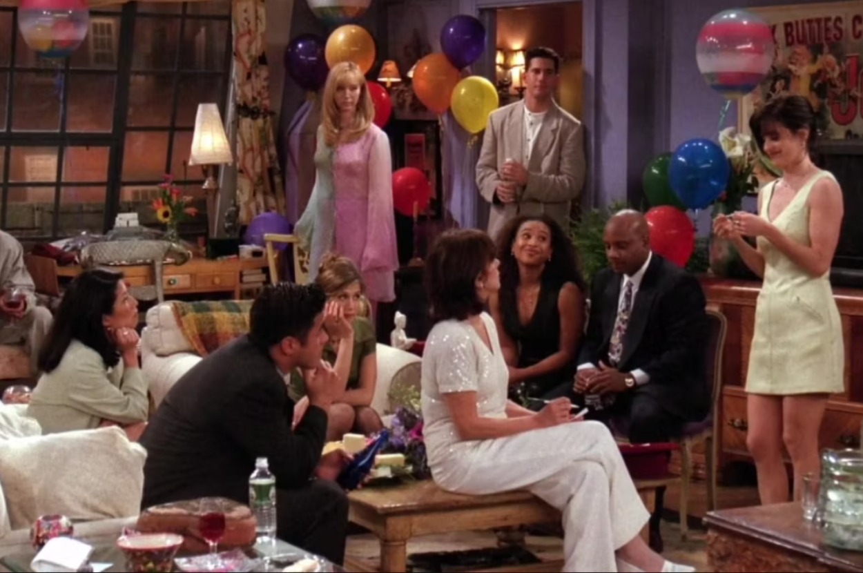 Group of people, including Monica, Rachel, Ross, Joey, Chandler, and Phoebe, gathered in a living room decorated with balloons, having a conversation