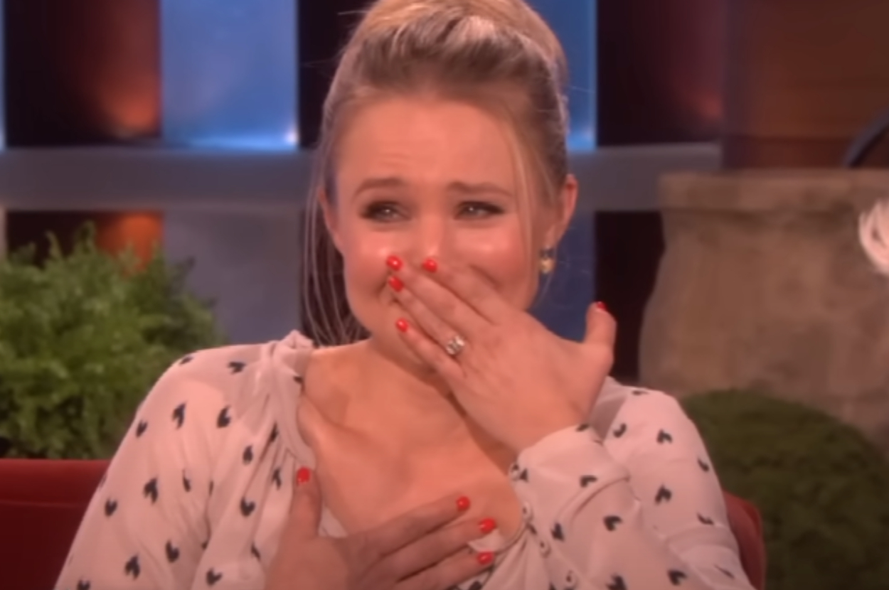 Kristen Bell on a talk show, wearing a patterned blouse with heart shapes, appears emotional with her hand over her mouth and chest
