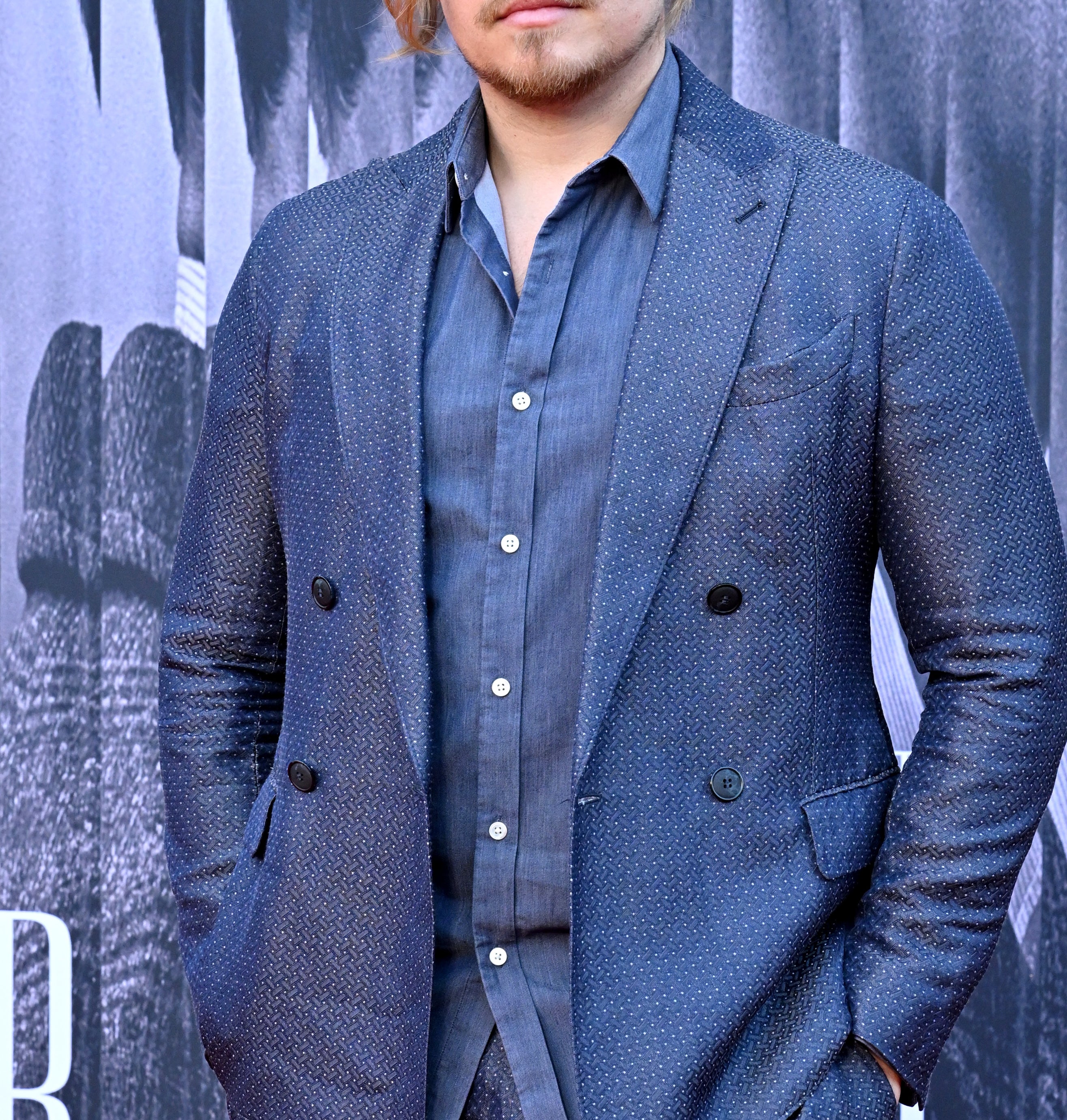 Dylan Sprouse standing on a red carpet, wearing a double-breasted suit with his hands in his pockets