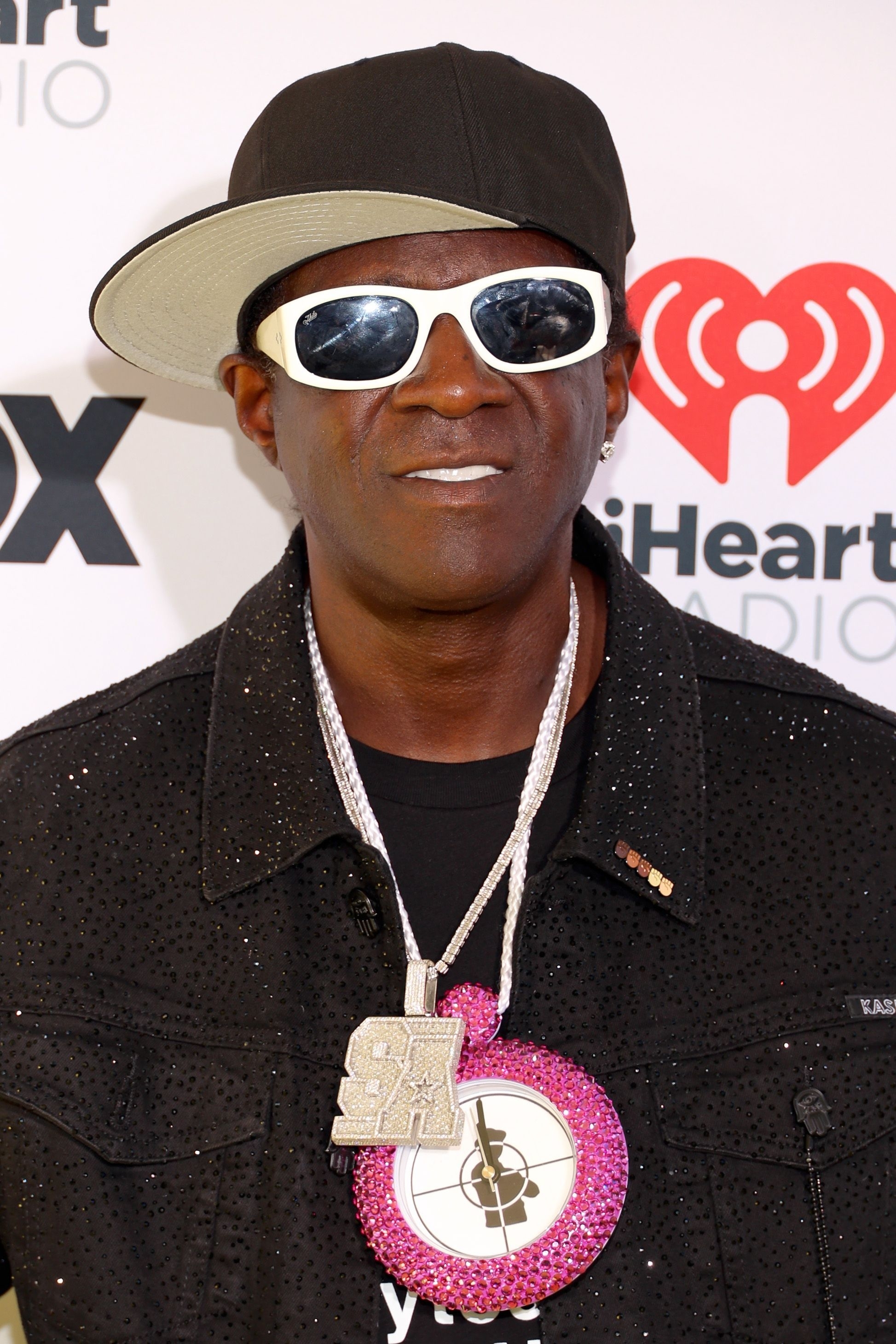 Flavor Flav at an iHeartRadio event, wearing a black sequined jacket, large clock necklace, white sunglasses, and a black cap