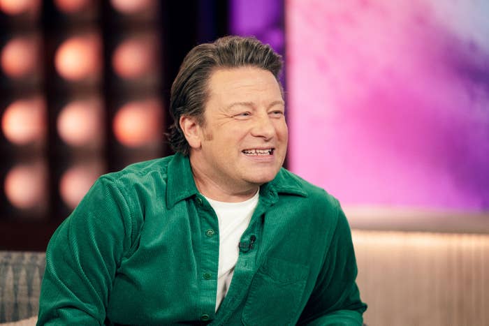 Jamie Oliver is seated, smiling, and wearing a button-up shirt over a white tee during an interview