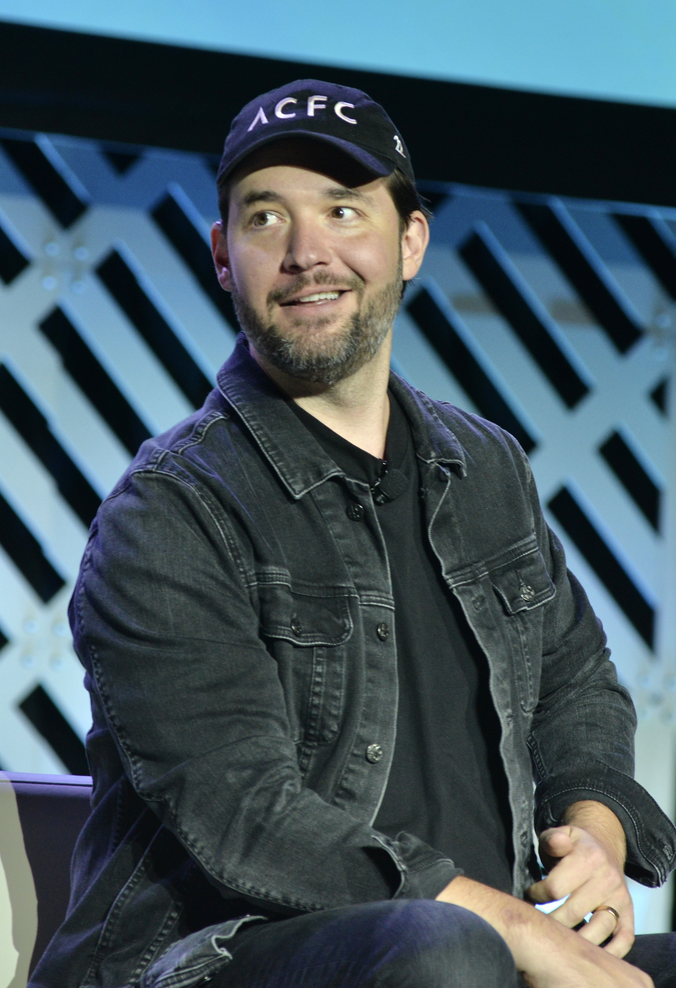 Alexis Ohanian sits on a stage wearing a black denim jacket, black shirt, and an ACFC cap, looking slightly to his left