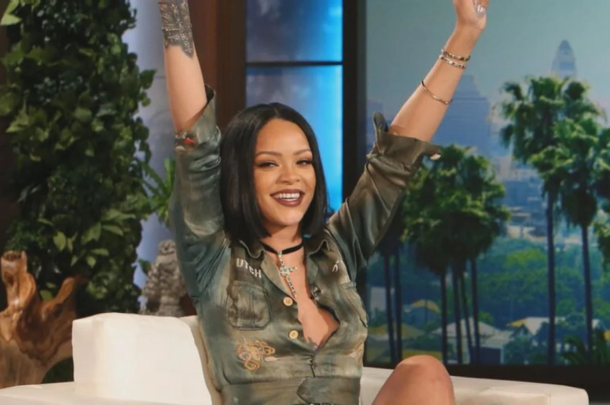 Rihanna raises her arms while seated on a talk show, wearing a stylish green patterned dress with short sleeves and a deep neckline