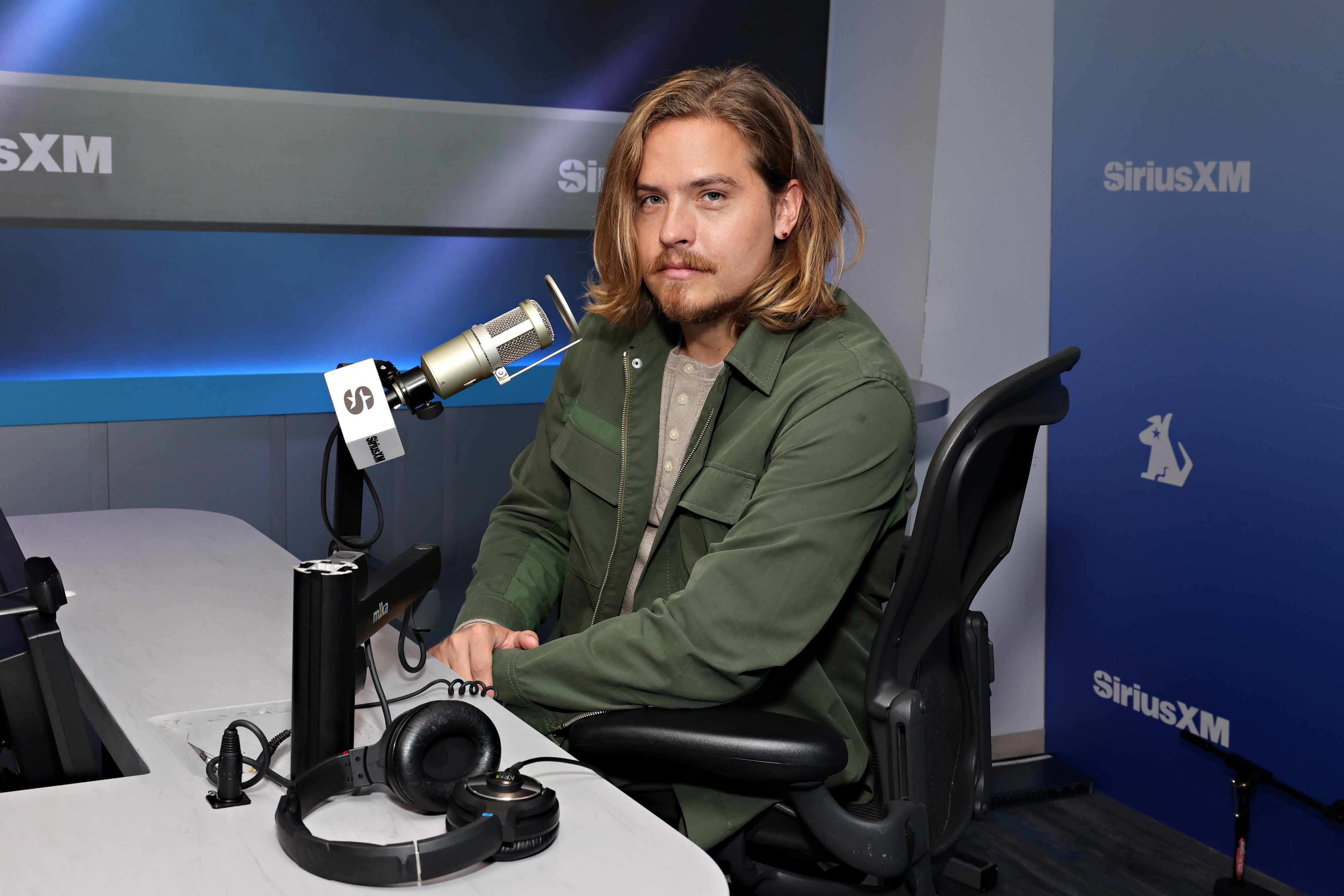 Dylan Sprouse seated at a SiriusXM studio, wearing a casual green jacket, posing with headphones and microphone equipment