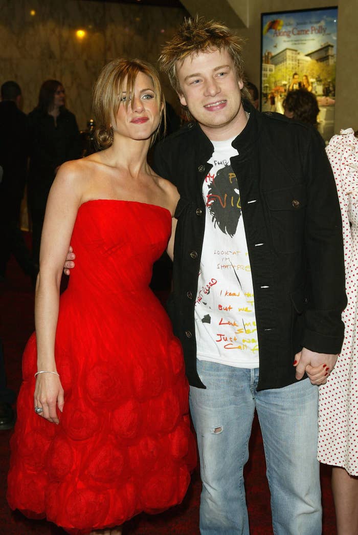 Jennifer Aniston in a strapless dress with floral details poses with Jamie Oliver in a graphic shirt and jeans at an event
