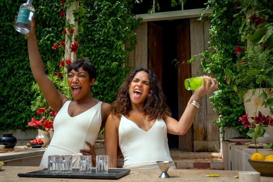 Tiffany Haddish and Rosario Dawson are celebrating and toasting with drinks, wearing white outfits in an outdoor setting adorned with greenery and flowers