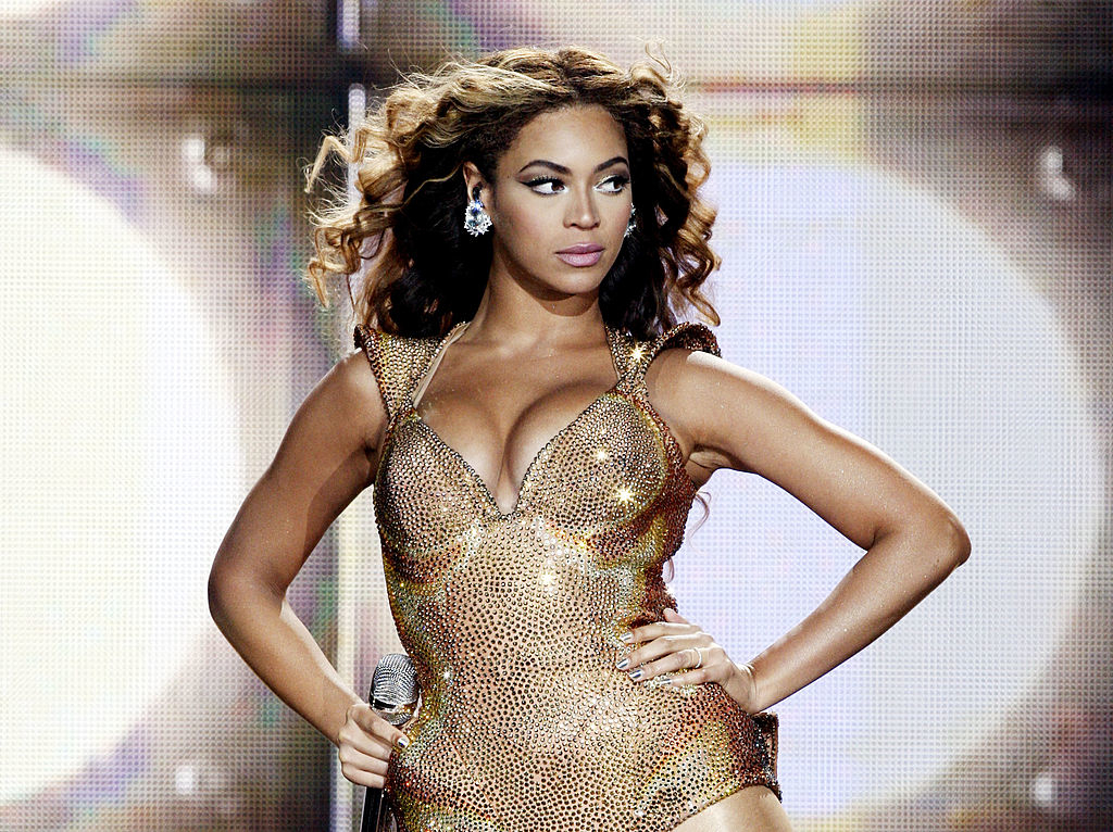 Beyoncé dressed in a shimmering, form-fitting bodysuit with structured shoulders, performing on stage, holding a microphone