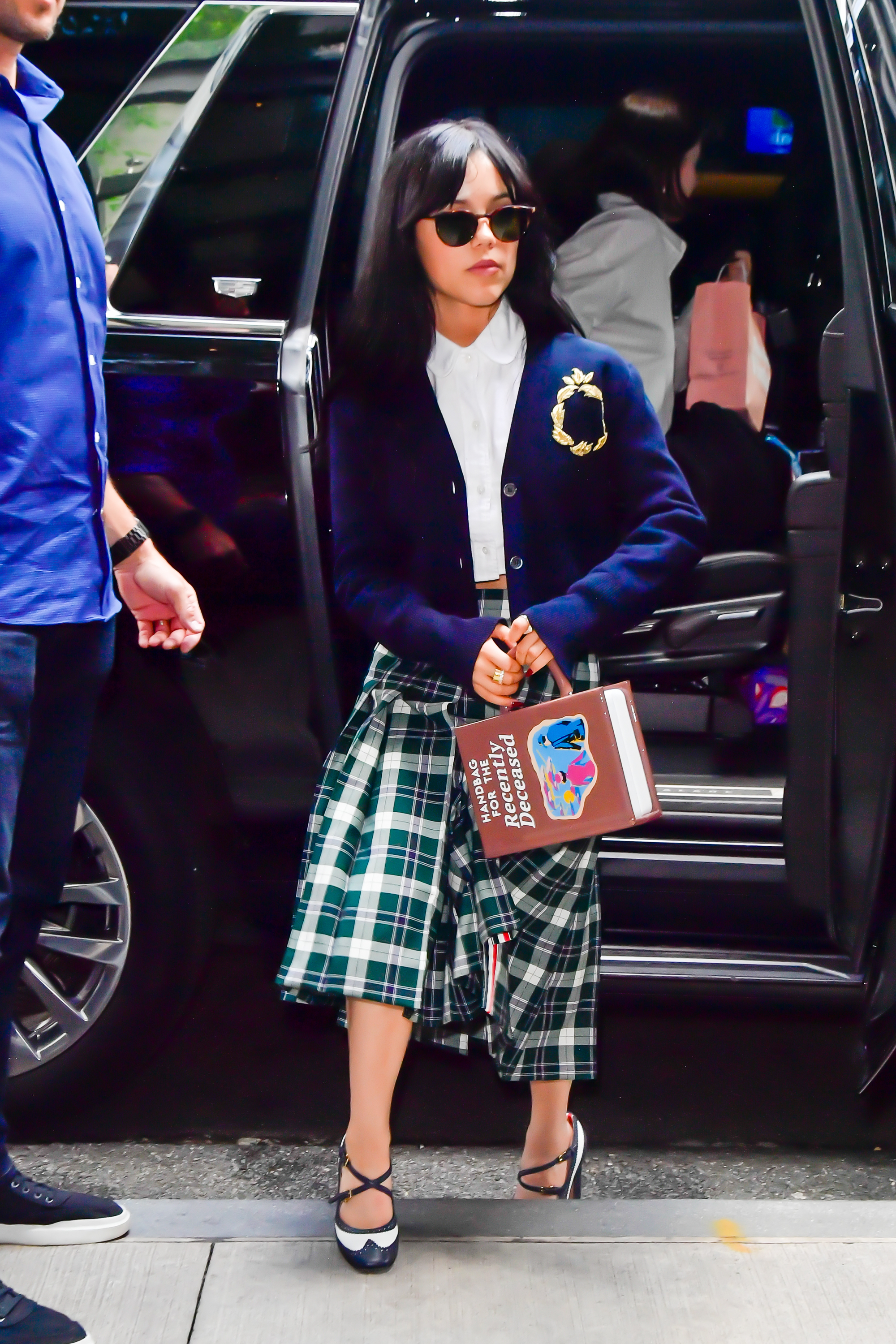 Jenna Ortega steps out of a car wearing a plaid skirt, white blouse, dark cardigan, sunglasses, and holding a lunchbox purse