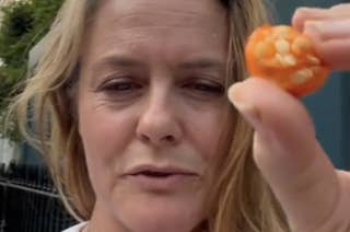 Alicia Silverstone holds up a small orange fruit while standing outside in front of a building