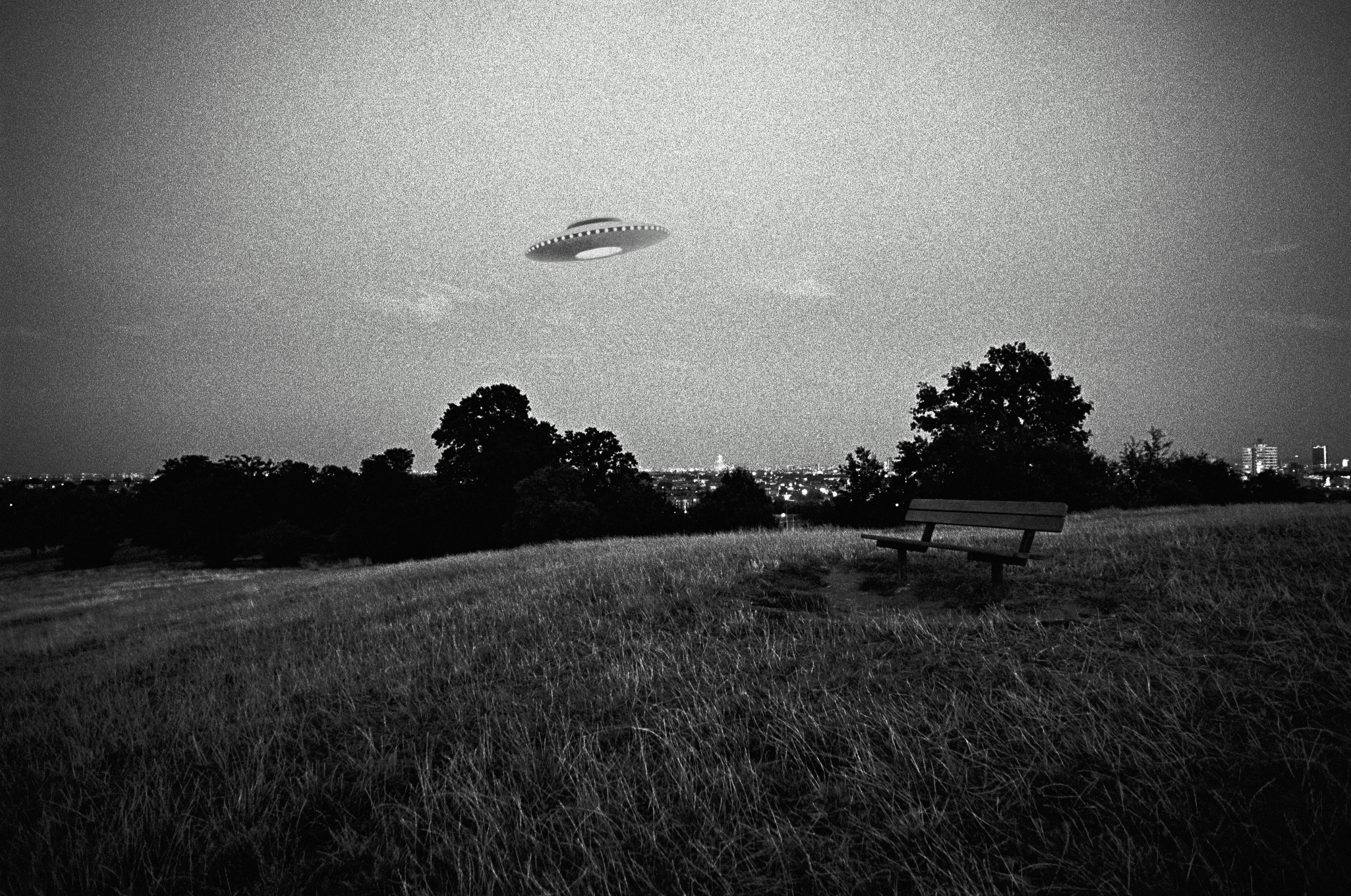 A UFO hovers in the sky over a grassy hill with a lone bench. City lights are visible in the distance under a darkened sky