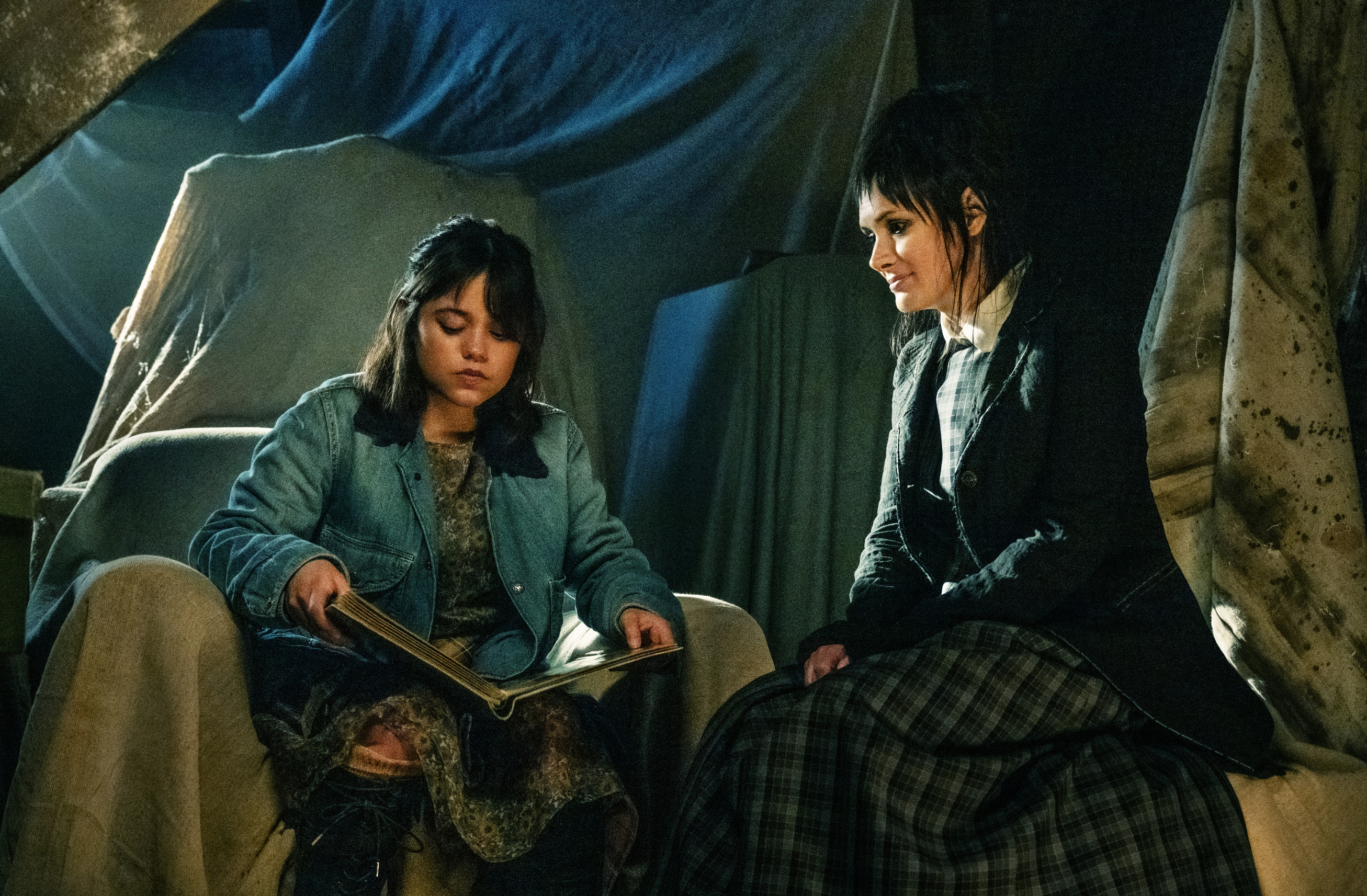 Jenna Ortega dressed casually while sitting and reading a book, with Christina Ricci wearing a suit jacket and plaid skirt sitting beside her, both in a dimly lit room