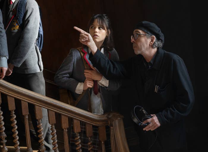 Jenna Ortega in school uniform looks on as Tim Burton, wearing black with a beret, points at something on a staircase