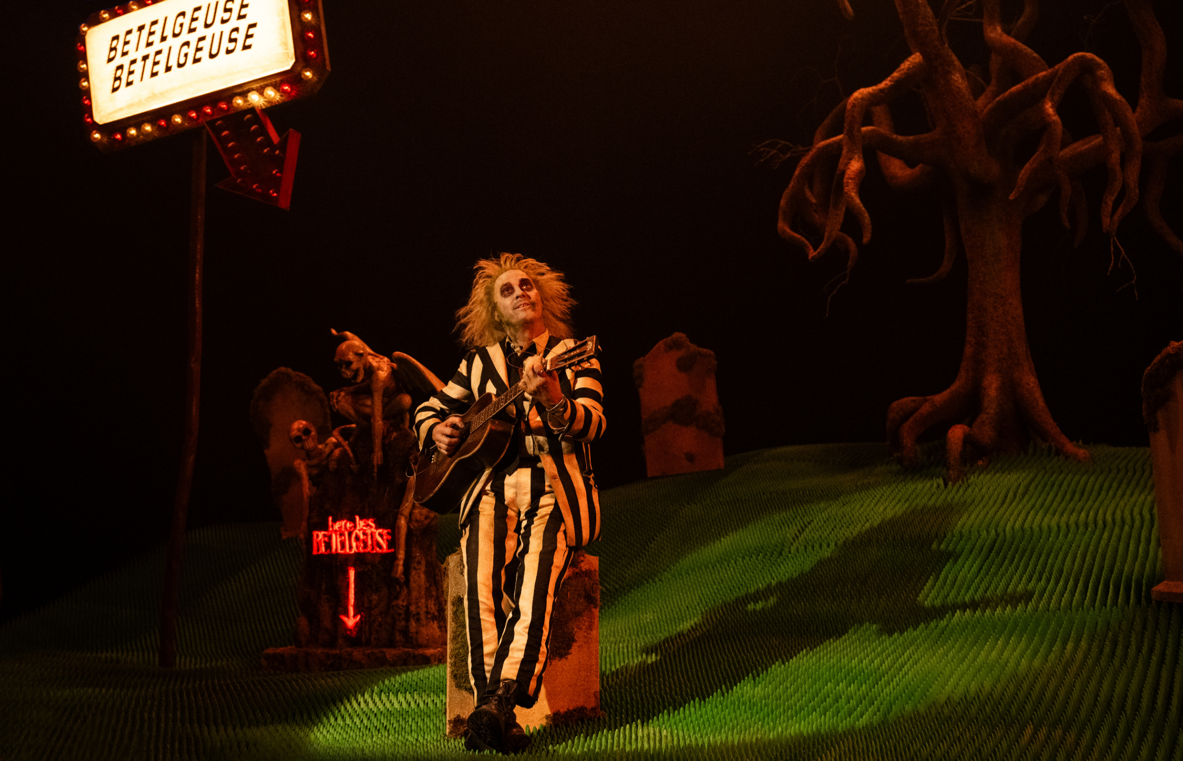 A performer dressed as Beetlejuice, in a black-and-white striped suit, plays a guitar on a stage with a graveyard set