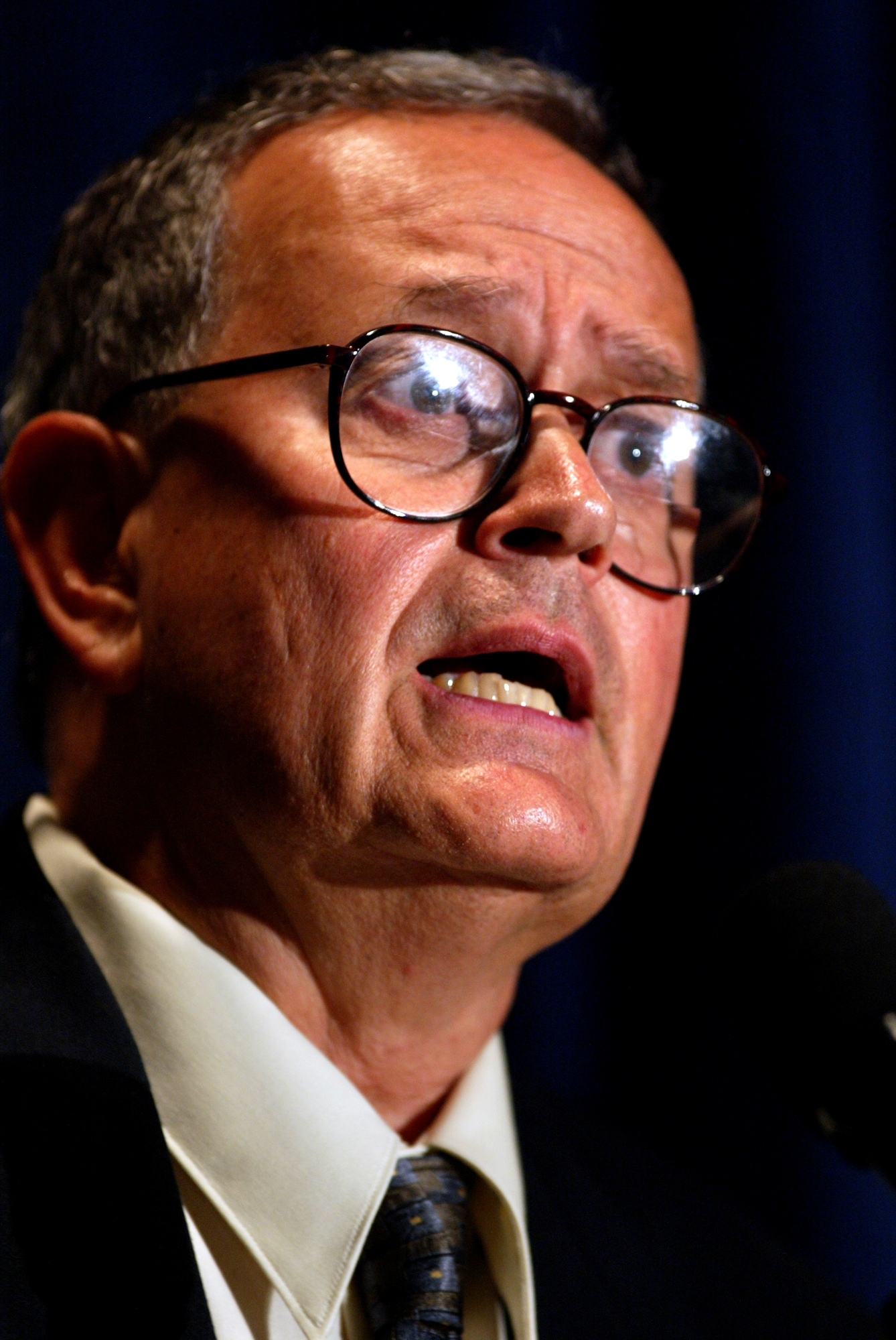 Camejo wearing glasses and a suit is speaking into a microphone at what appears to be a formal event or press conference