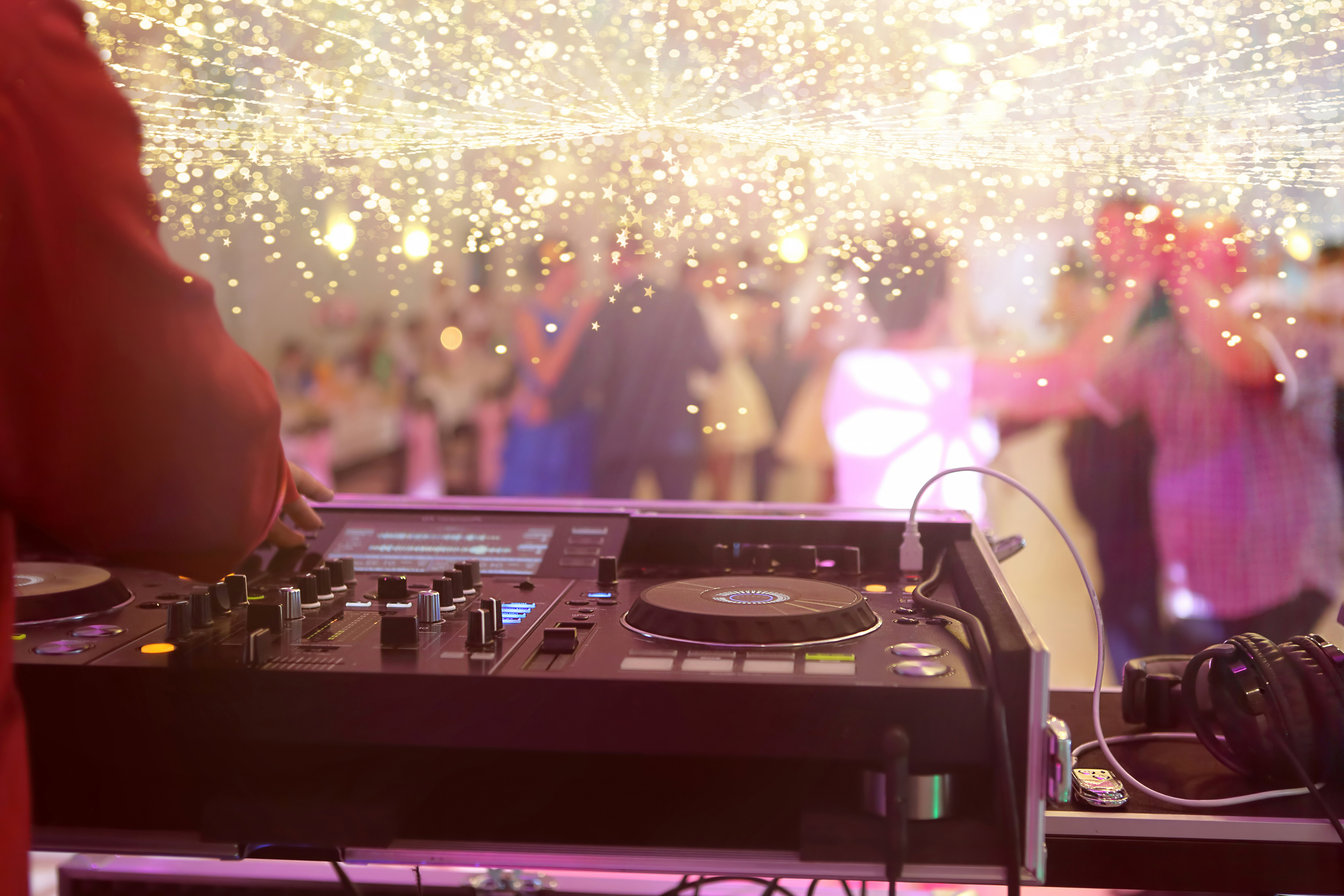 A DJ performs at an event, operating a DJ console with lights and a crowd of people dancing in the background