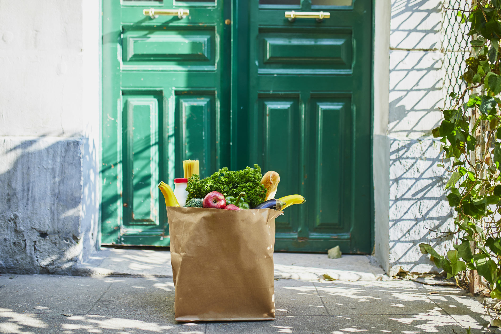 Paper grocery bag filled with fresh produce, pasta, and a baguette placed on a doorstep in front of a double green door