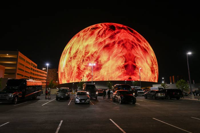 A giant spherical structure in a parking lot displays dynamic fire-like visuals on its surface at night. Several vehicles are parked around it