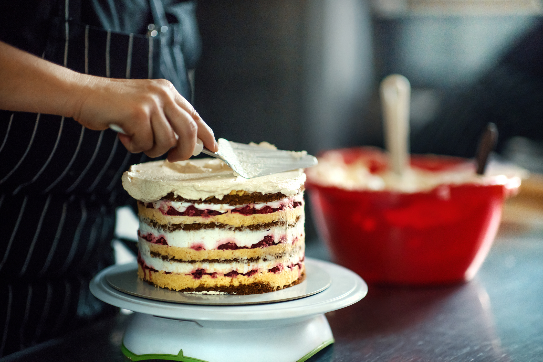 Close-up of a person in a striped apron spreading frosting on a layer cake with visible cream and fruit filling. A red mixing bowl is in the background