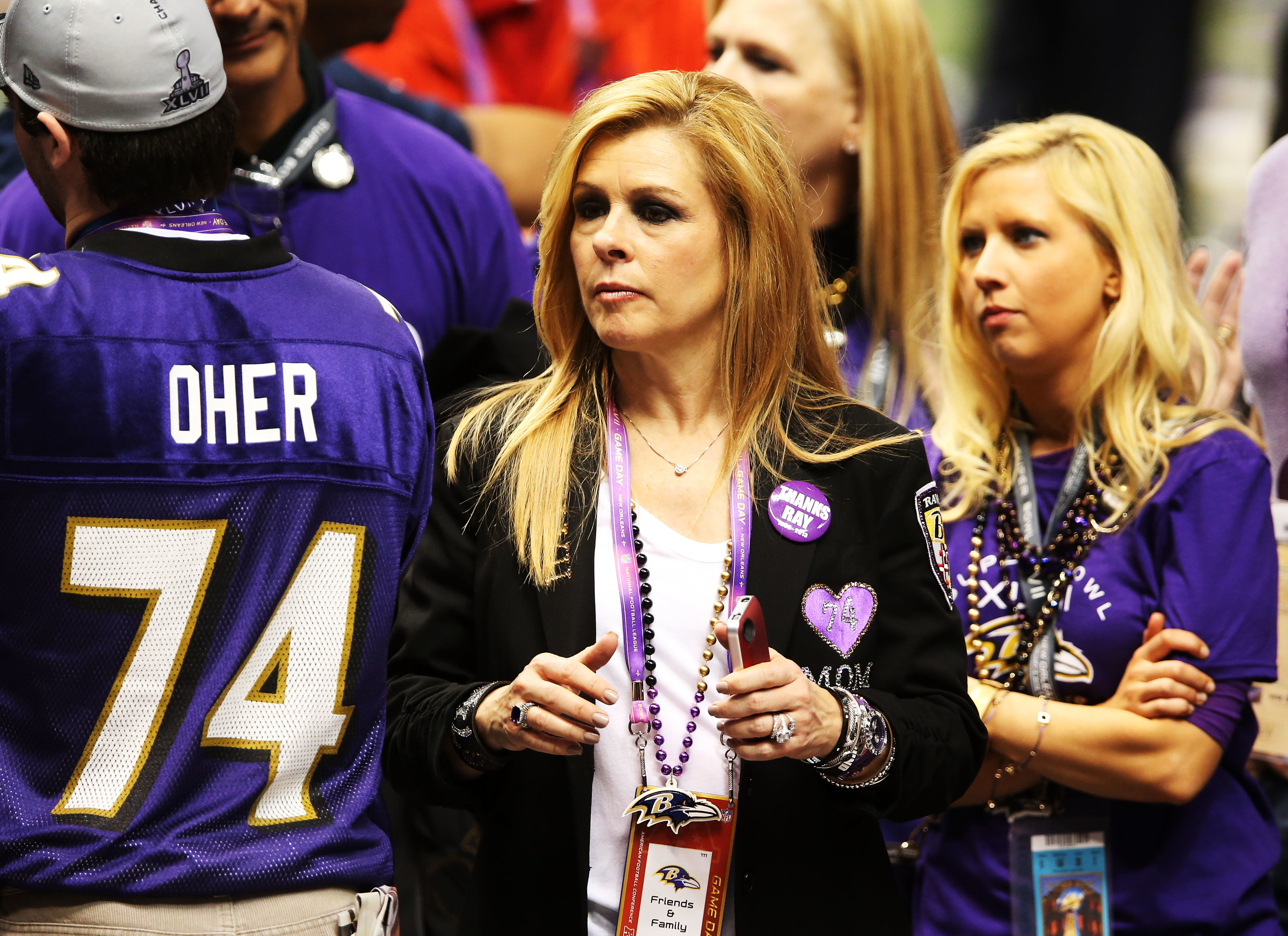 A group of people, including Leigh Anne Tuohy, wearing Baltimore Ravens attire at a public event. She holds a drink and a Ravens lanyard, while others chat nearby