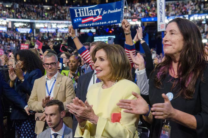 Hillary Clinton supporters cheer at an event. A woman in a yellow dress, possibly Nancy Pelosi, stands in the foreground. Signs for Hillary Clinton for President are visible