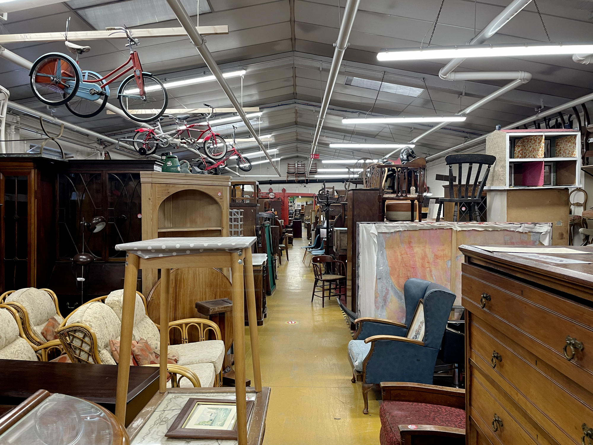 A large furniture warehouse filled with various types of used and antique furniture, including chairs, tables, cabinets, and shelves, with bicycles hanging from the ceiling