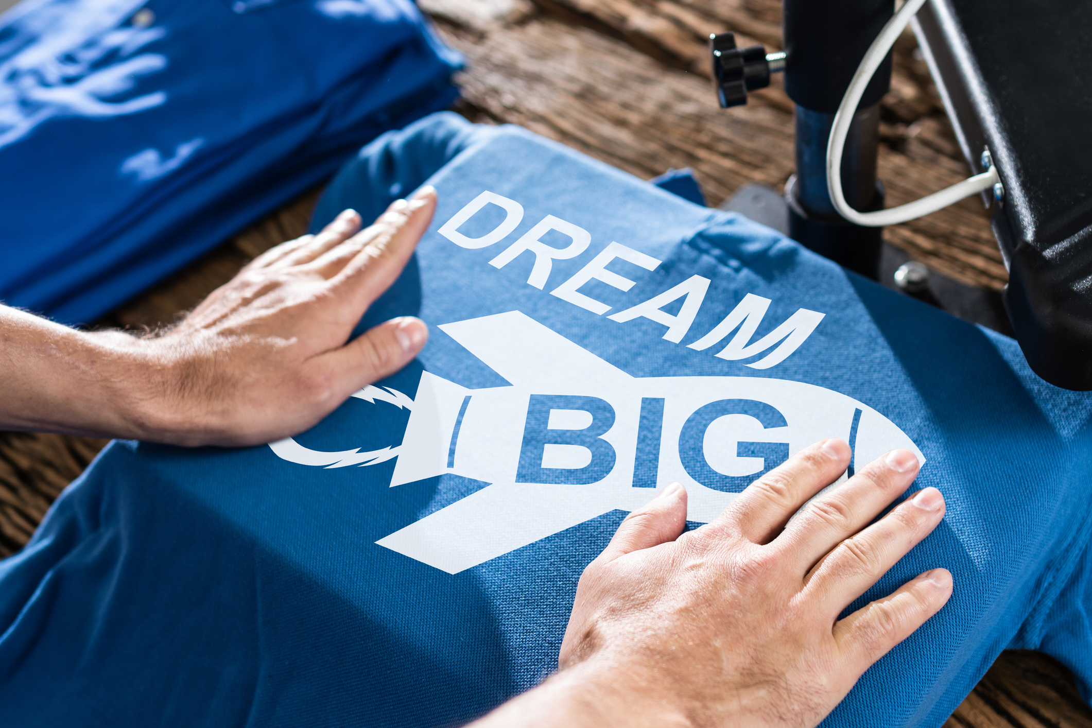 Close-up of hands using a heat press to print "Dream Big" with a rocket graphic on a shirt, symbolizing ambition and motivation