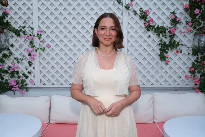 Maya Rudolph stands in front of a lattice backdrop with flowers, wearing a light dress and a cardigan, smiling