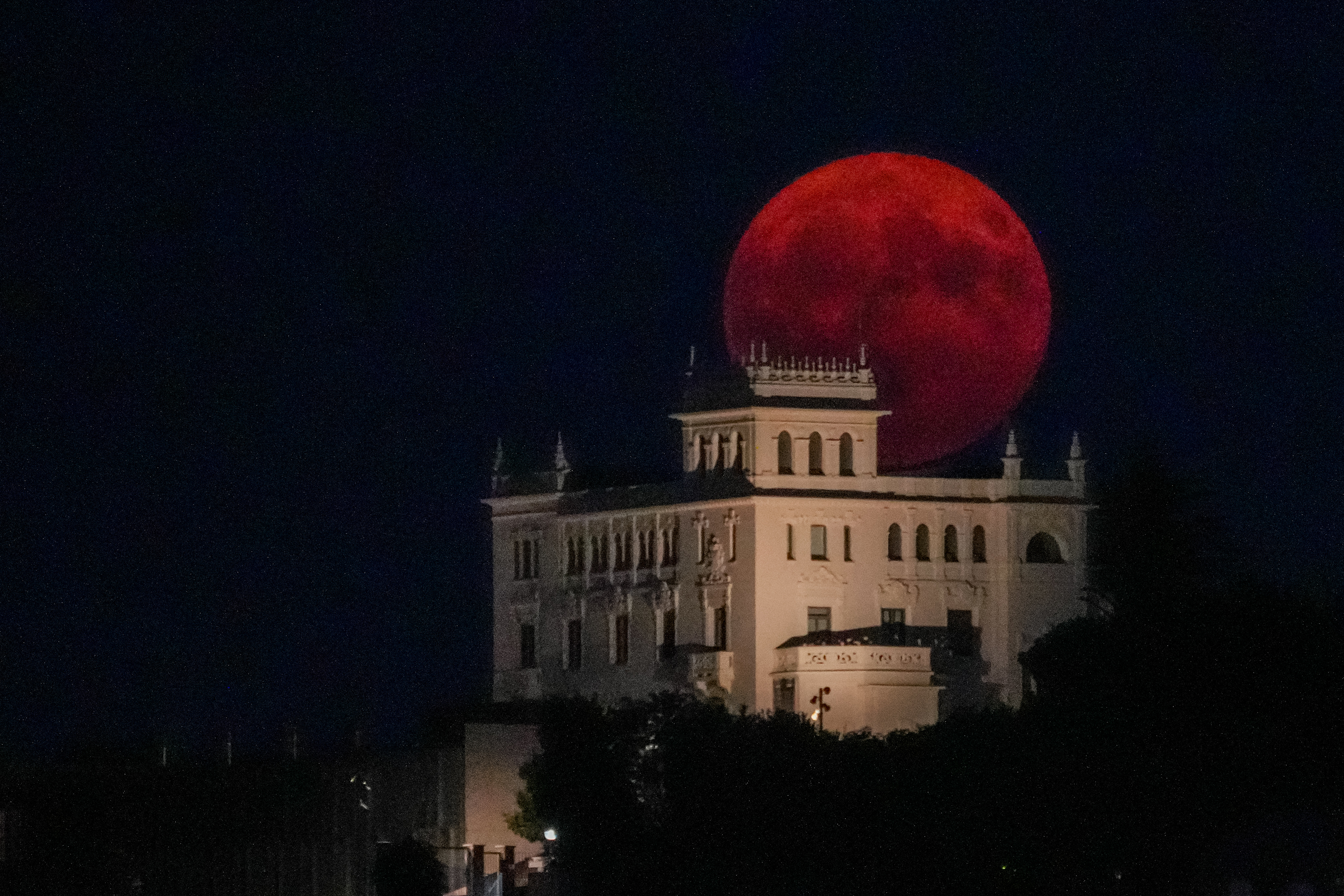 A large, full red moon rises behind a historic, white castle-like building against a dark sky