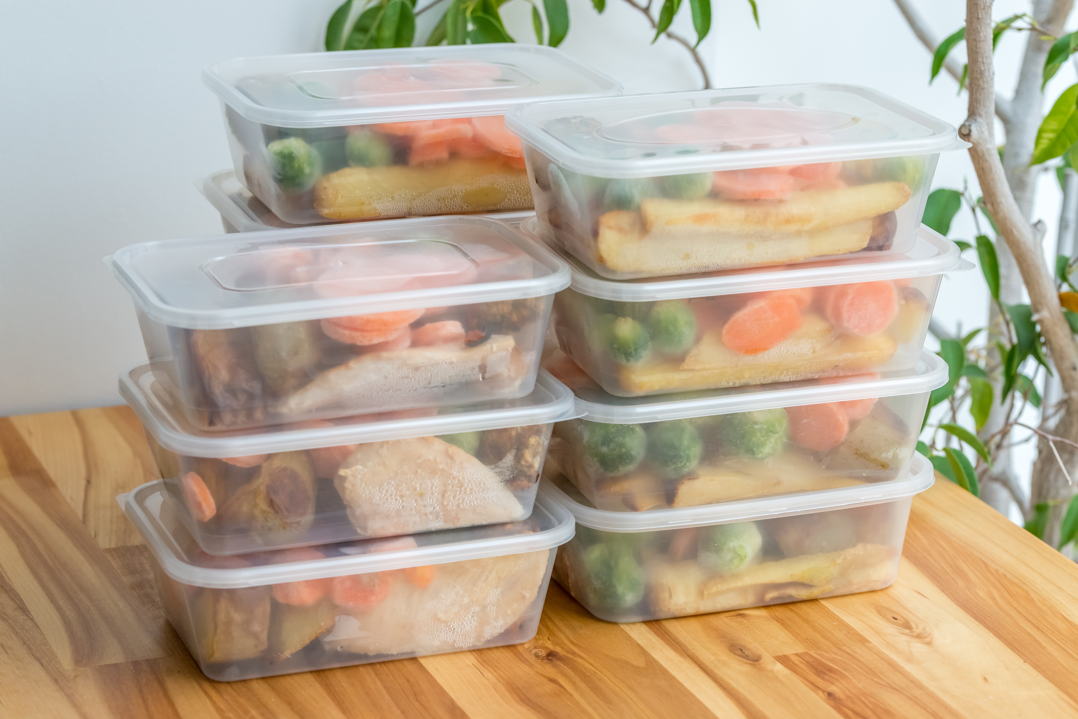 Stacked plastic containers filled with assorted meal prepped vegetables and proteins on a wooden table, with a plant in the background