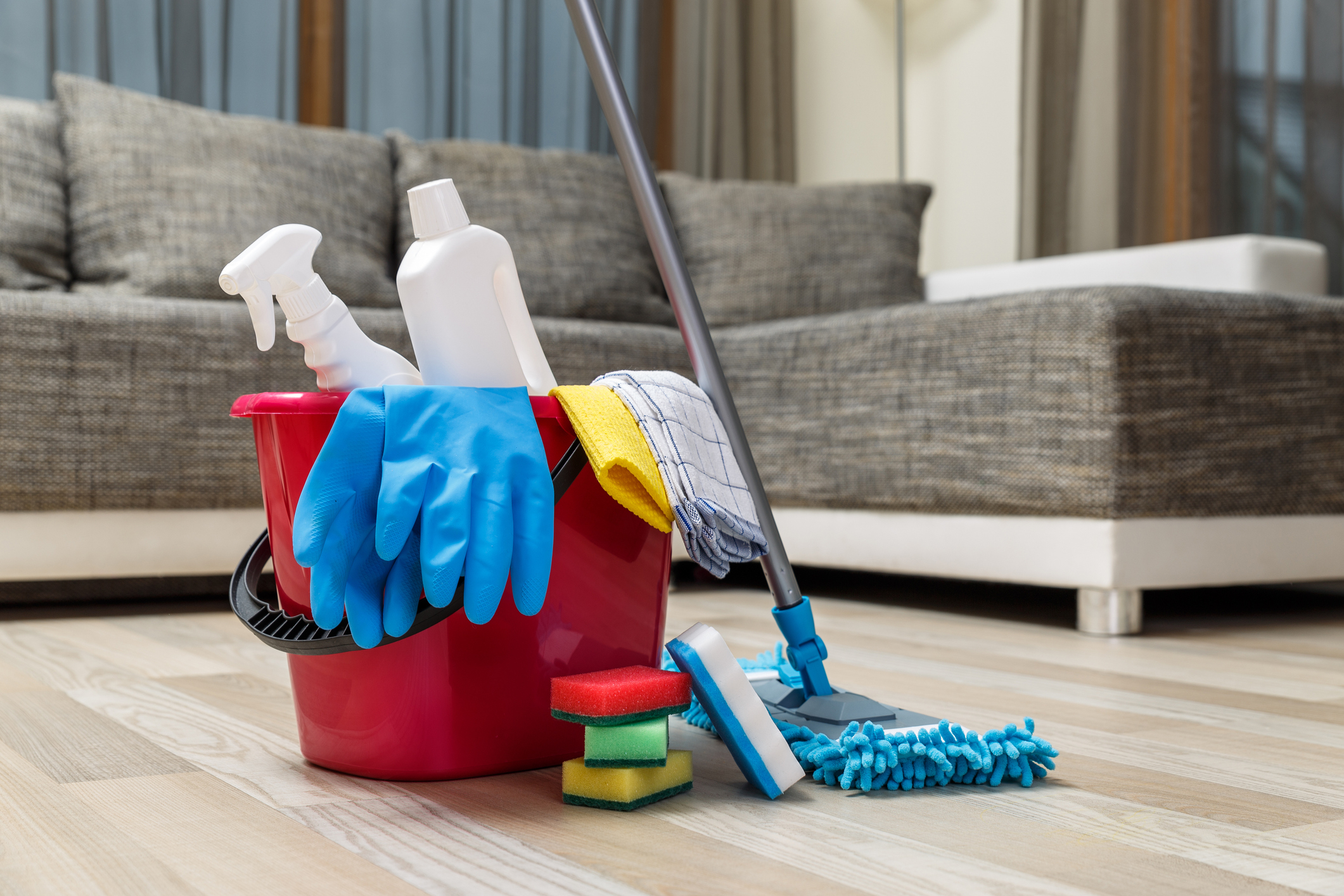 Cleaning supplies including gloves, sponges, spray bottles, and a mop in a red bucket on a hardwood floor with a couch in the background