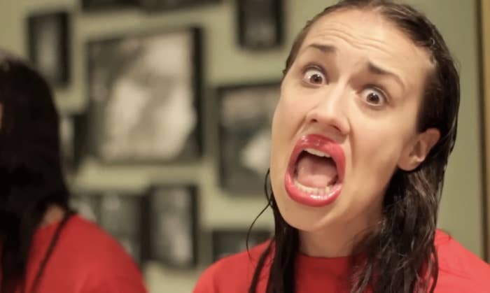 Colleen Ballinger, as her character Miranda Sings, wears exaggerated red lipstick and open-mouthed expression, stands in front of a wall of framed pictures