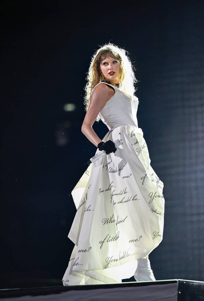 Taylor Swift performs on stage wearing a long dress adorned with handwritten lyrics, striking a dramatic pose with curly hair and a confident expression