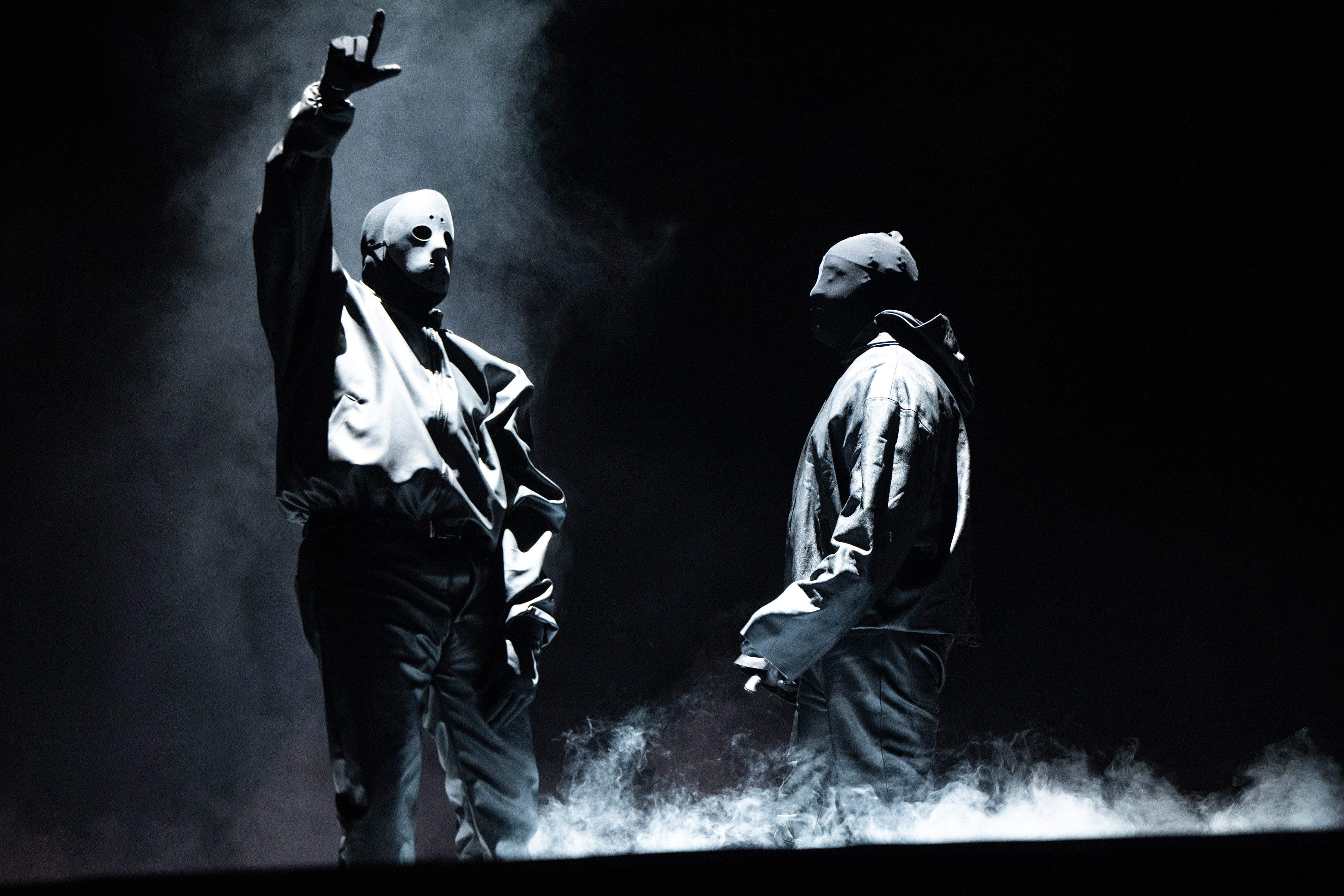 Ye and Ty Dolla $ign on stage with dramatic lighting and smoke effects. One performer gestures with their hand while the other stands facing them