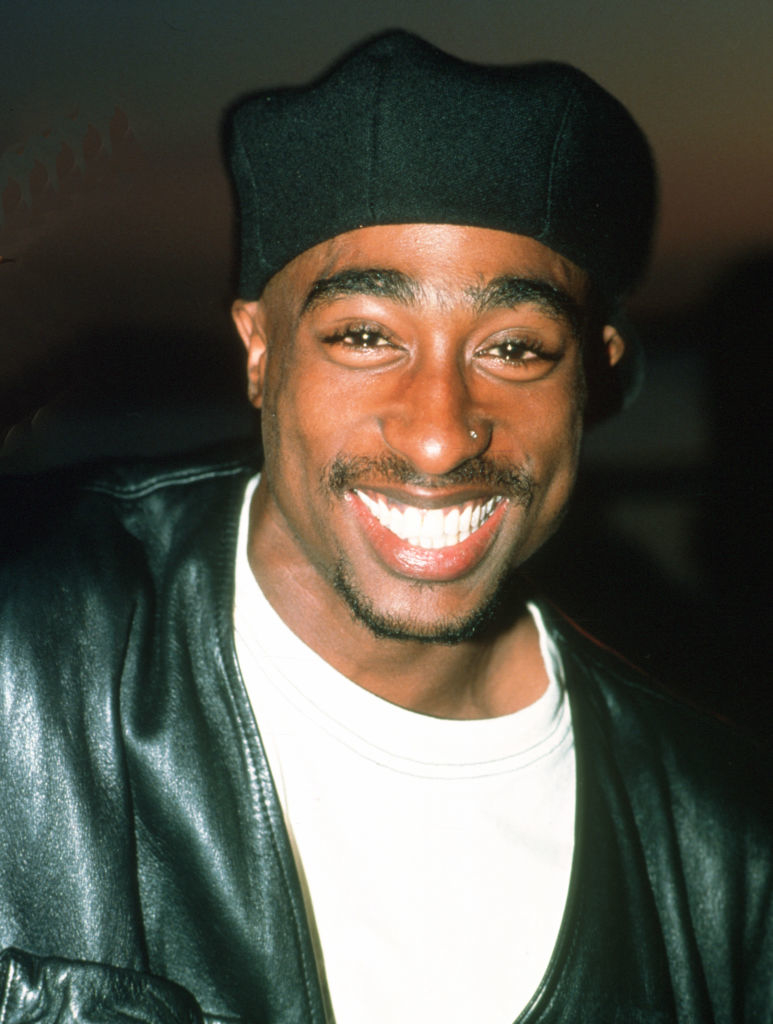 Tupac Shakur smiling broadly, wearing a black beret, a white shirt, and a black leather jacket