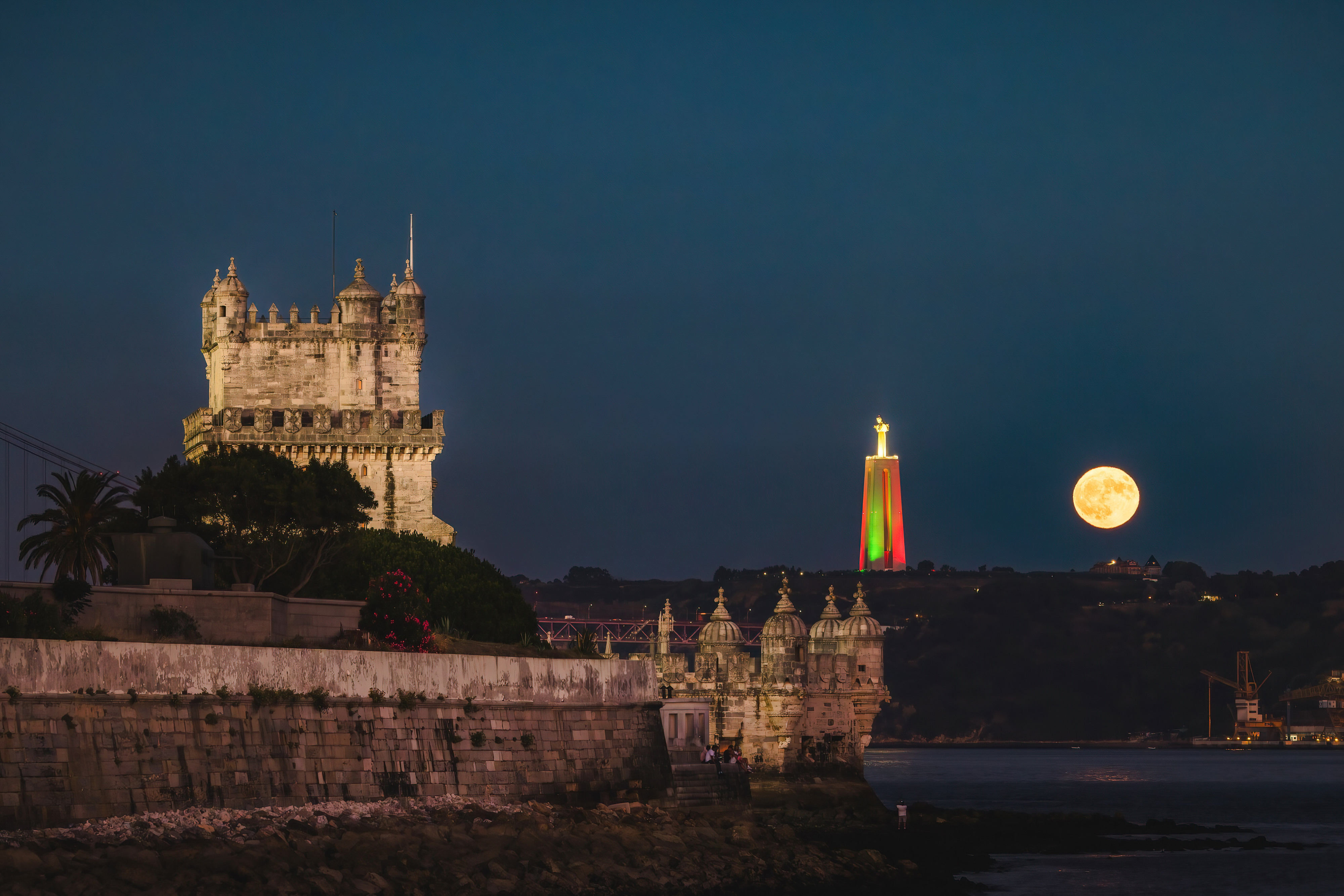 A night view showing the Belém Tower and the Sanctuary of Christ the King statue illuminated with a full moon in Lisbon, Portugal