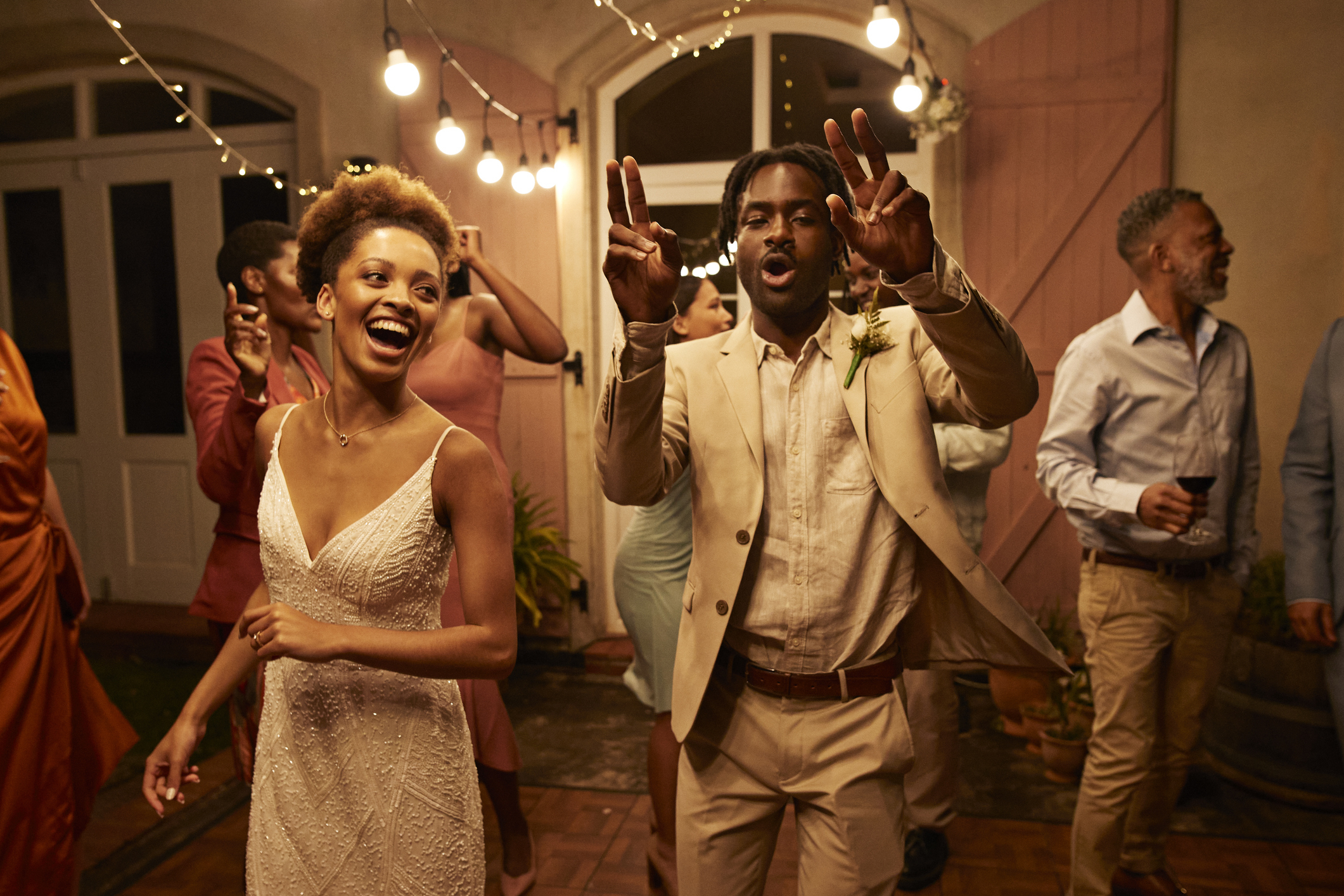 People dancing and celebrating at a party; a woman in a sleeveless dress and a man in a beige suit are in the foreground, other guests are in the background