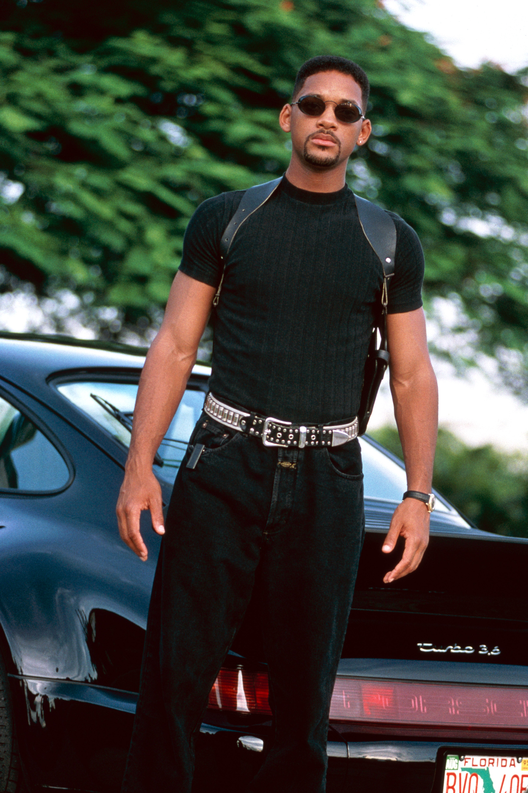 Will Smith standing confidently in front of a Porsche. He is wearing sunglasses, a black T-shirt, and black pants with a studded belt