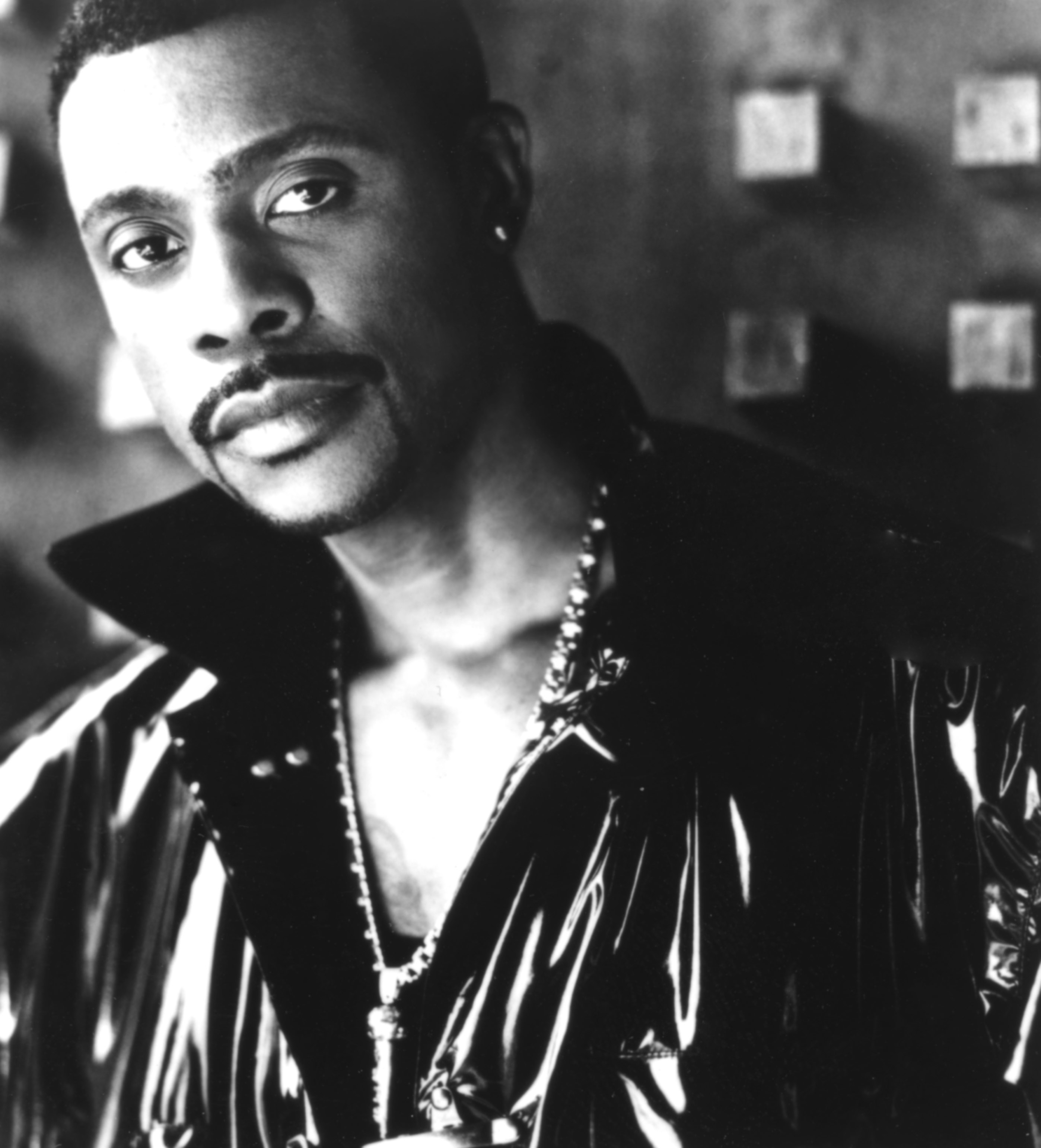 Keith Sweat wears a shiny jacket and chain necklace, posing against a backdrop