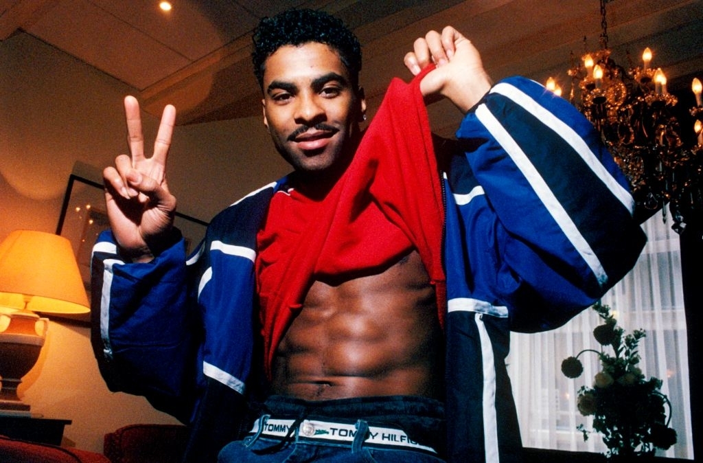 Man holding up his shirt to show his abs while smiling and flashing a peace sign indoors. He is wearing a Tommy Hilfiger belt and jacket