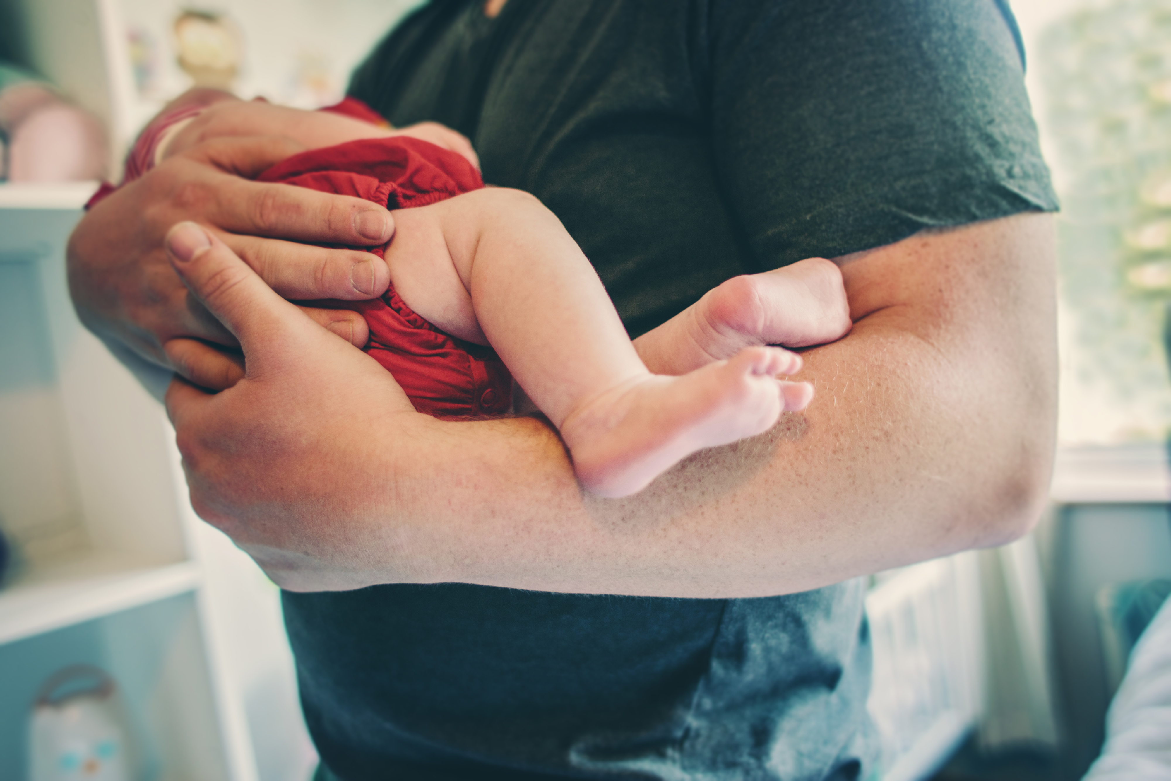 A person in a t-shirt cradles a baby wearing a onesie, with the baby’s legs and feet visible. Their surroundings suggest an indoor setting