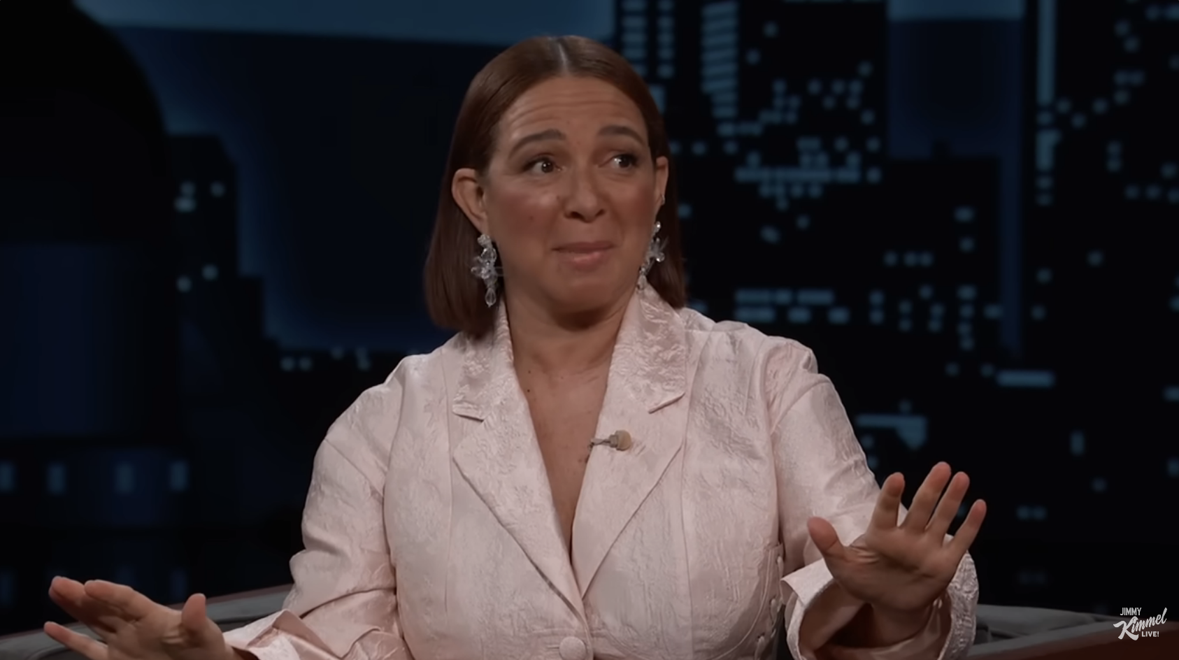 Maya Rudolph is on a TV show set, wearing a floral patterned blazer and statement earrings, gesturing with both hands