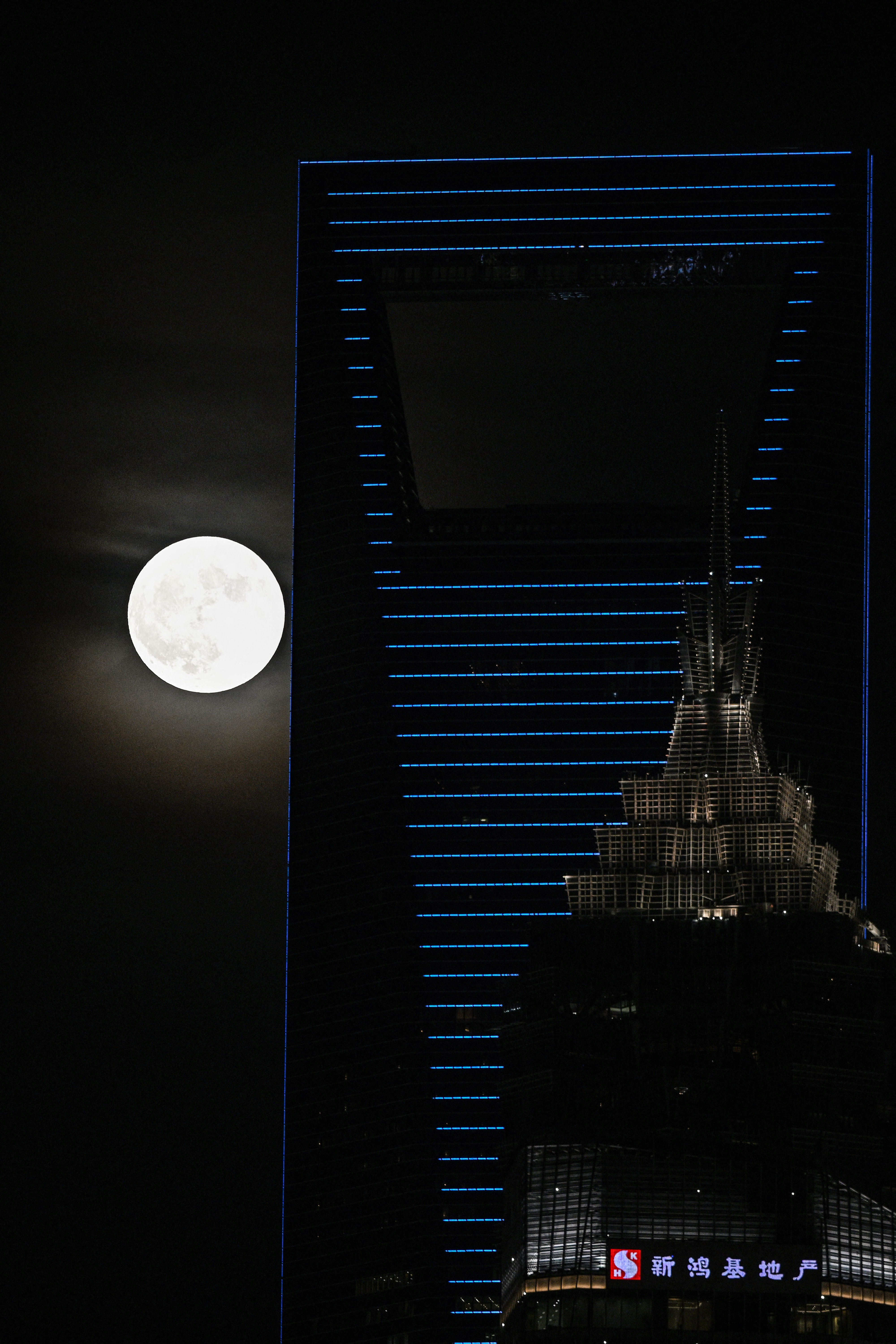 Full moon near the top of a modern skyscraper at night, with another illuminated building partially visible in the foreground