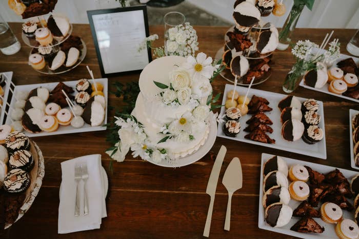 A beautifully arranged dessert table featuring a white-tiered cake adorned with white flowers, various pastries, and cutlery on a wooden surface