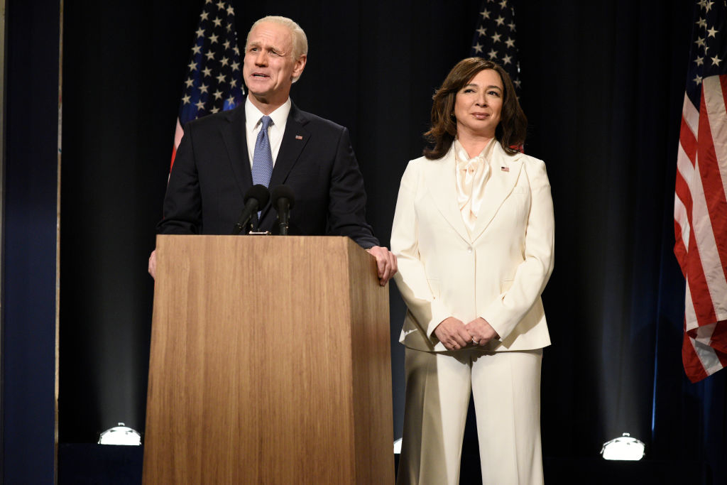 Joe Biden, in a suit and tie, stands at a podium with Maya Rudolph, dressed in a white suit, on stage with U.S. flags in the background
