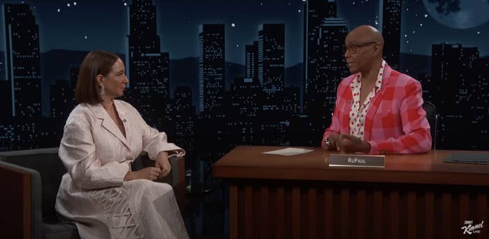 Maya Rudolph and RuPaul having a conversation on a talk show set, with a city skyline backdrop. RuPaul wears a pink checkered jacket, and Maya wears a white dress