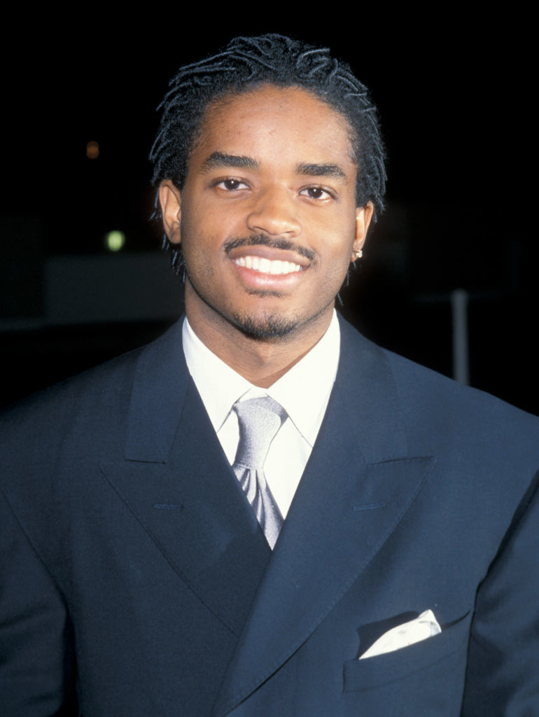 André 3000 is smiling and wearing a double-breasted suit with a tie and pocket square