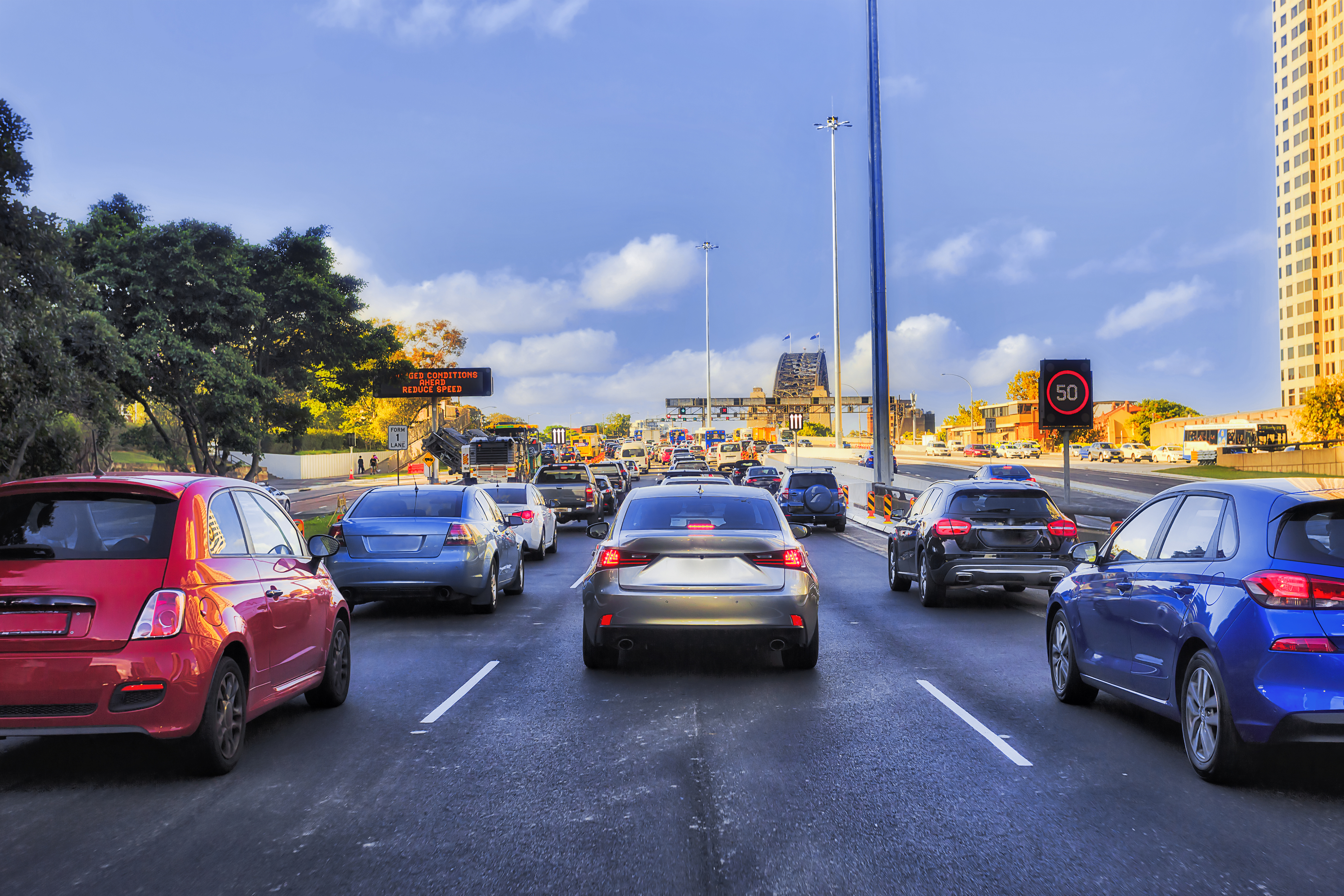 Dense traffic endures on a highway with a speed limit of 50 displayed. Numerous vehicles are in several lanes, including two red cars on the left and right